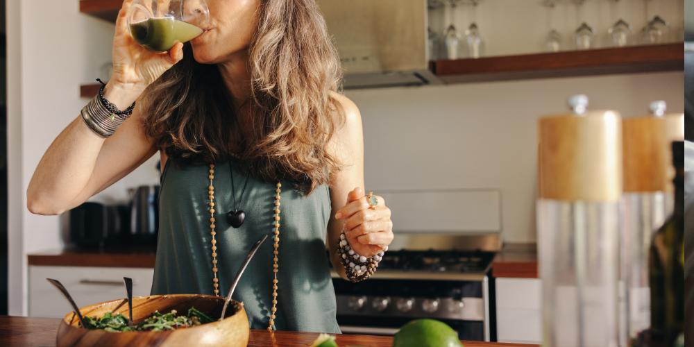 Kobieta w średnim wieku pijąca zielony koktajl w swojej kuchni, obok miski z warzywną sałatką – przykład zdrowych nawyków wspierających metabolizm i dobre samopoczucie w menopauzie.