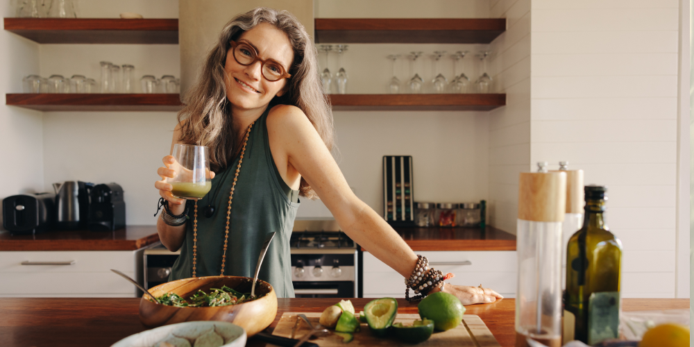 Kobieta w średnim wieku w kuchni, uśmiechnięta, z zielonym koktajlem w dłoni, przy stole z awokado i zdrową sałatką – symbol zdrowego stylu życia i diety wspierającej organizm w okresie menopauzy.